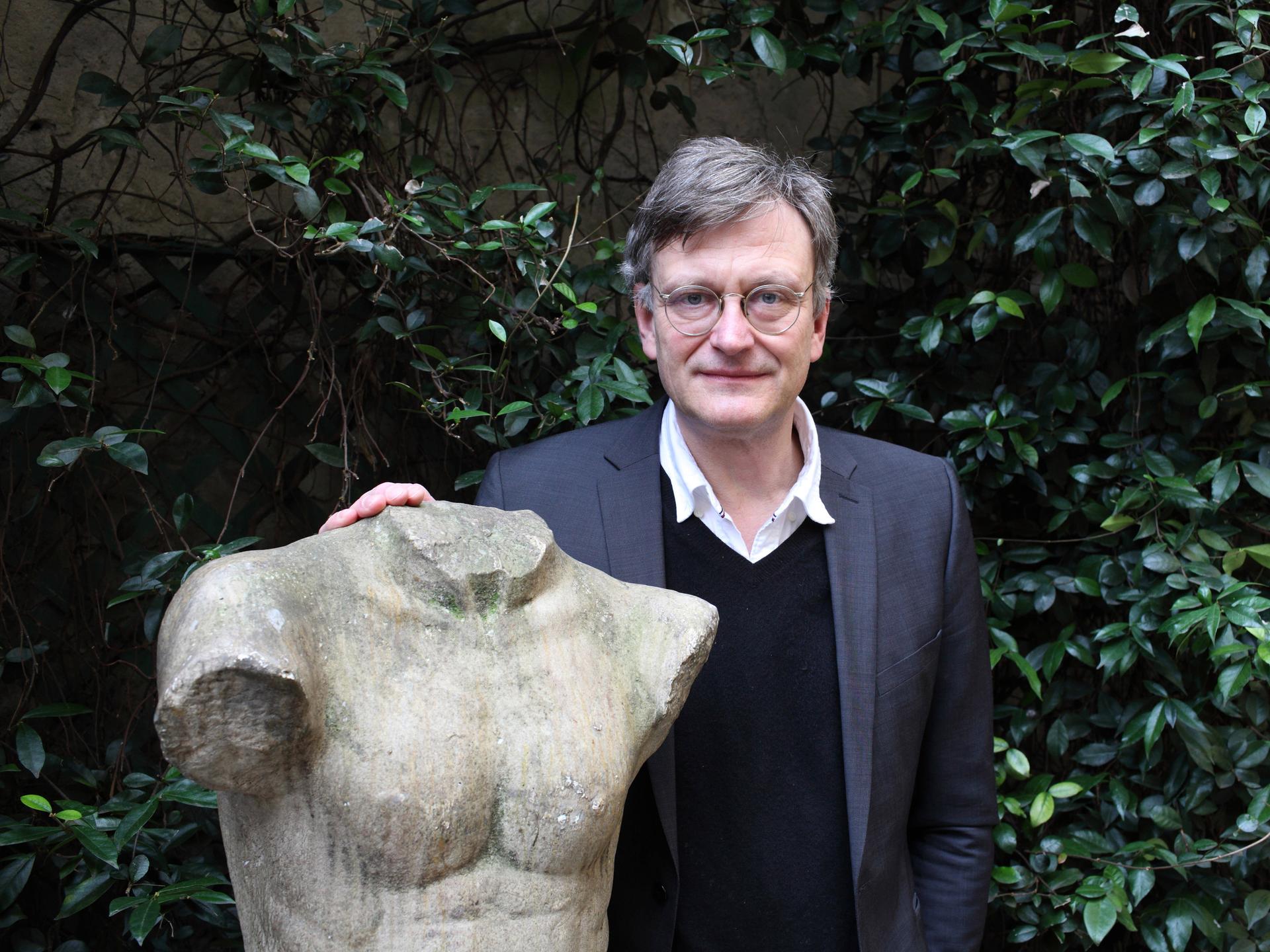 Hans von Trotha steht in einem Garten an eine Statue gelehnt, die einen Männerkörper mit abgeschlagenem Kopf und Gliedmaßen zeigt. Hans von Trotha steht in einem Garten an eine Statue gelehnt, die einen Männerkörper mit abgeschlagenem Kopf und Gliedmaßen zeigt.