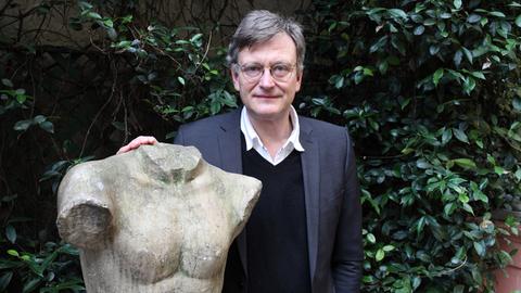 Hans von Trotha steht in einem Garten an eine Statue gelehnt, die einen Männerkörper mit abgeschlagenem Kopf und Gliedmaßen zeigt. Hans von Trotha steht in einem Garten an eine Statue gelehnt, die einen Männerkörper mit abgeschlagenem Kopf und Gliedmaßen zeigt.