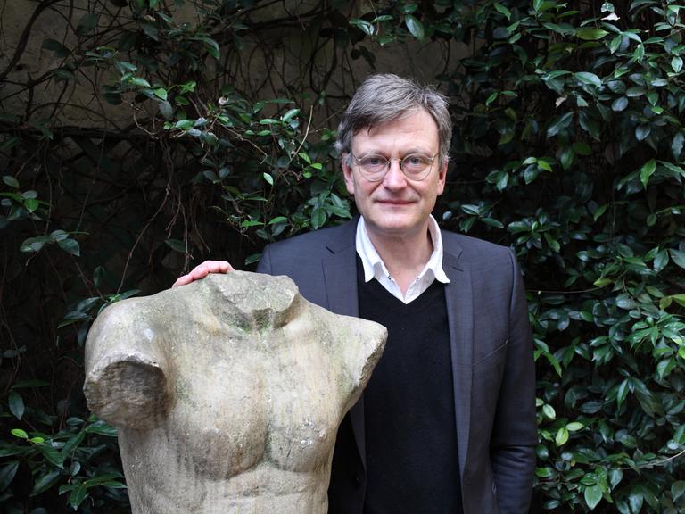 Hans von Trotha steht in einem Garten an eine Statue gelehnt, die einen Männerkörper mit abgeschlagenem Kopf und Gliedmaßen zeigt. Hans von Trotha steht in einem Garten an eine Statue gelehnt, die einen Männerkörper mit abgeschlagenem Kopf und Gliedmaßen zeigt.