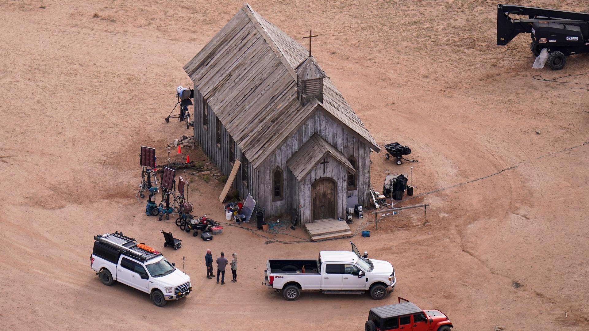 Ein historisch wirkendes, hölzernes Kirchengebäude auf rotem Sand, davor moderne geländegängige Fahrzeuge