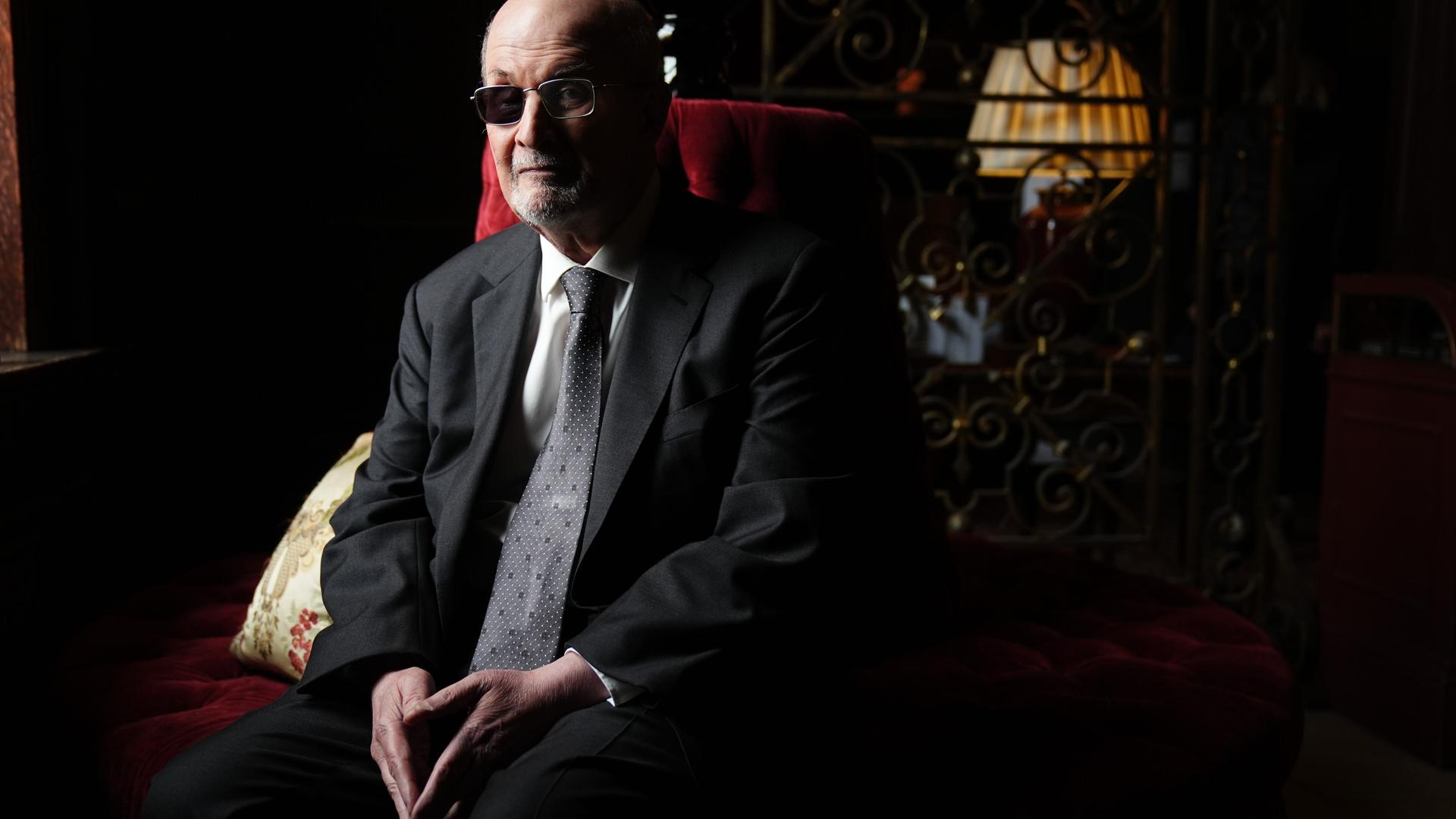 Der Autor Salman Rushdie sitzt im Halbdunkel auf einem roten Sofa und blickt ernst in die Kamera, die Hände im Schoß.