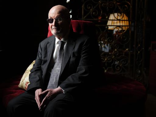 Der Autor Salman Rushdie sitzt im Halbdunkel auf einem roten Sofa und blickt ernst in die Kamera, die Hände im Schoß. Der Autor Salman Rushdie sitzt im Halbdunkel auf einem roten Sofa und blickt ernst in die Kamera, die Hände im Schoß.