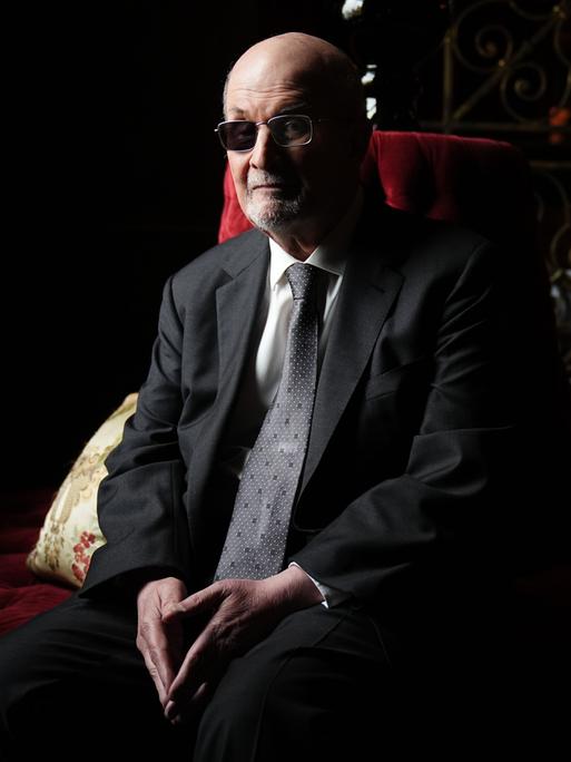 Der Autor Salman Rushdie sitzt im Halbdunkel auf einem roten Sofa und blickt ernst in die Kamera, die Hände im Schoß.