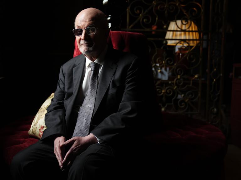 Der Autor Salman Rushdie sitzt im Halbdunkel auf einem roten Sofa und blickt ernst in die Kamera, die Hände im Schoß.