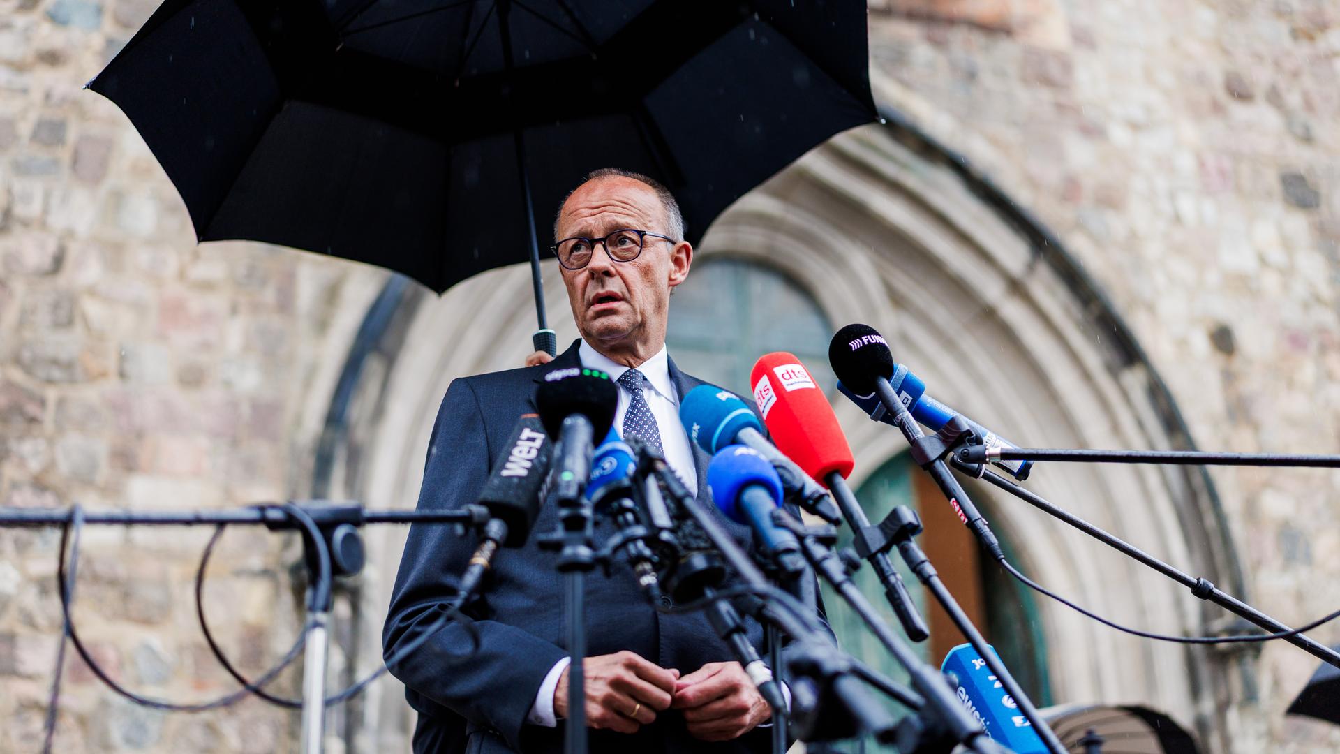 Friedrich Merz steht an zahlreichen Mikrofonen und gibt ein Statement ab. Über ihm ist ein großer schwarzer Regenschirm zu sehen. Friedrich Merz steht an zahlreichen Mikrofonen und gibt ein Statement ab. Über ihm ist ein großer schwarzer Regenschirm zu sehen.