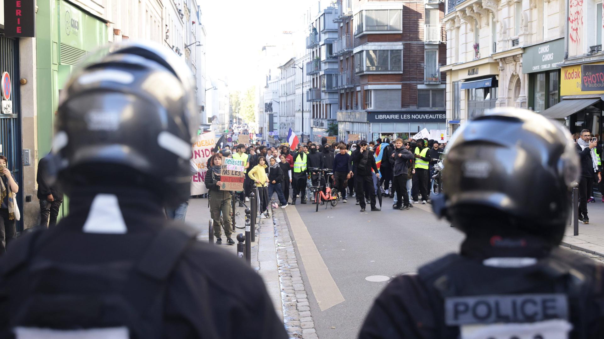 Demonstrierende ziehen durch eine Straße in Paris; vorne im Bild sind zwei Polizisten von hinten zu sehen.
      