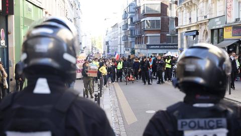 Demonstrierende ziehen durch eine Straße in Paris; vorne im Bild sind zwei Polizisten von hinten zu sehen.
      