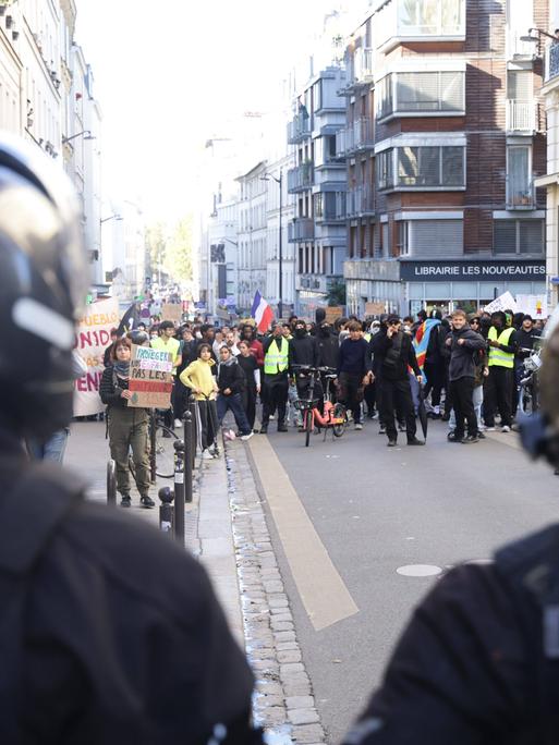 Demonstrierende ziehen durch eine Straße in Paris; vorne im Bild sind zwei Polizisten von hinten zu sehen.
      