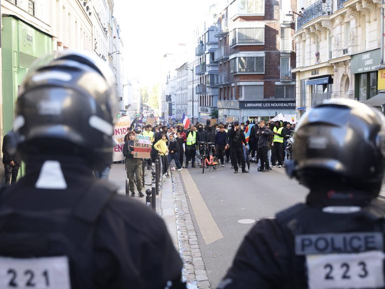 Demonstrierende ziehen durch eine Straße in Paris; vorne im Bild sind zwei Polizisten von hinten zu sehen.
      