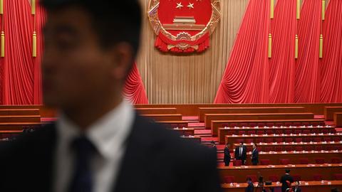 Blick in den Plenarsaal des Nationalen Volkskongresses in China. Zwischen den Bänken stehen Menschen. An den Wänden hängen überdimensional große rote Flaggen.