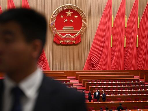 Blick in den Plenarsaal des Nationalen Volkskongresses in China. Zwischen den Bänken stehen Menschen. An den Wänden hängen überdimensional große rote Flaggen. Blick in den Plenarsaal des Nationalen Volkskongresses in China. Zwischen den Bänken stehen Menschen. An den Wänden hängen überdimensional große rote Flaggen.