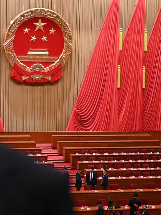 Blick in den Plenarsaal des Nationalen Volkskongresses in China. Zwischen den Bänken stehen Menschen. An den Wänden hängen überdimensional große rote Flaggen. Blick in den Plenarsaal des Nationalen Volkskongresses in China. Zwischen den Bänken stehen Menschen. An den Wänden hängen überdimensional große rote Flaggen.