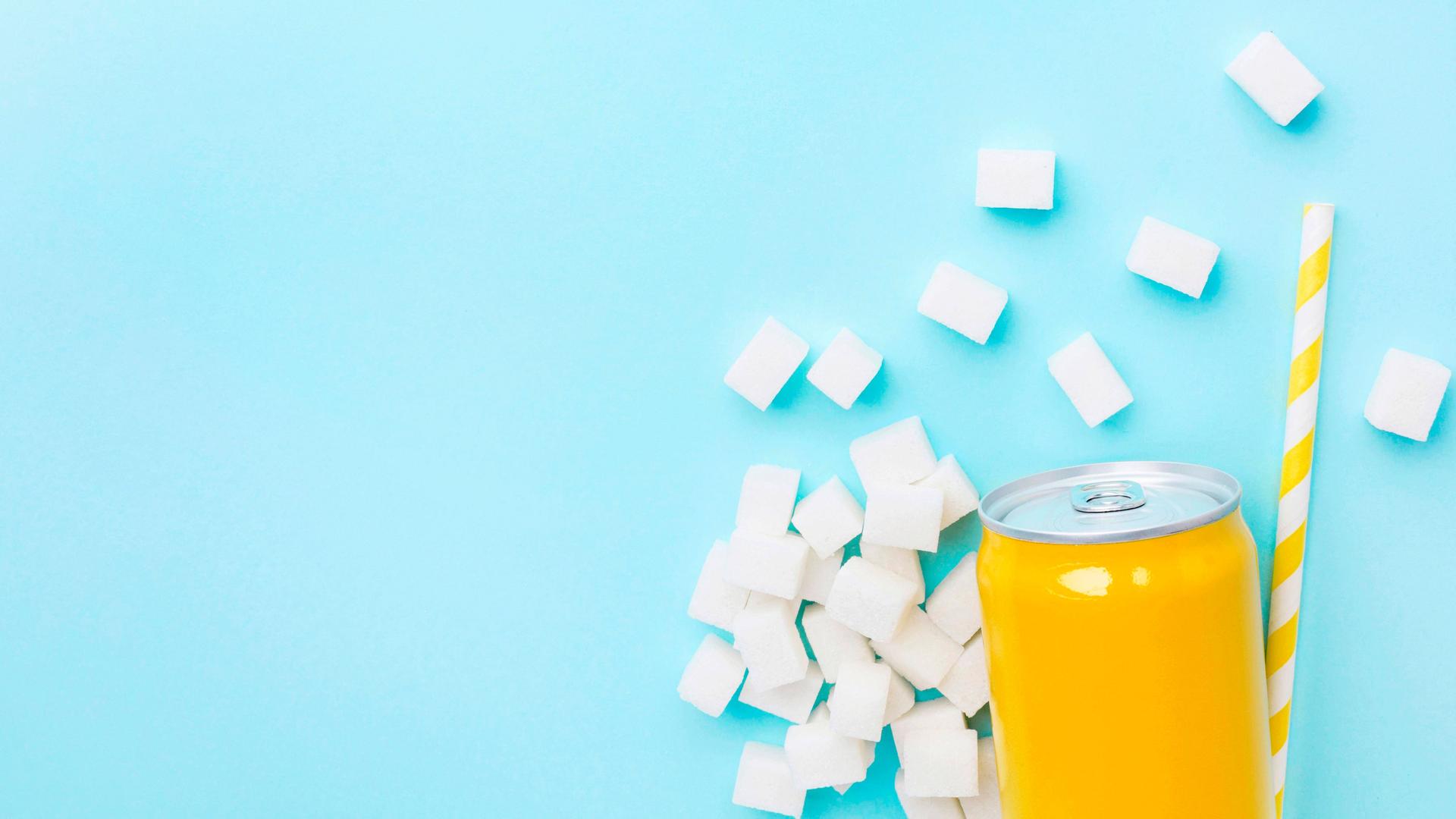 Gelbe Softdrinkdose auf hellblauen Hintergrund. Um die Dose herum liegen Würfelzucker und ein gelb-weißer Strohhalm.