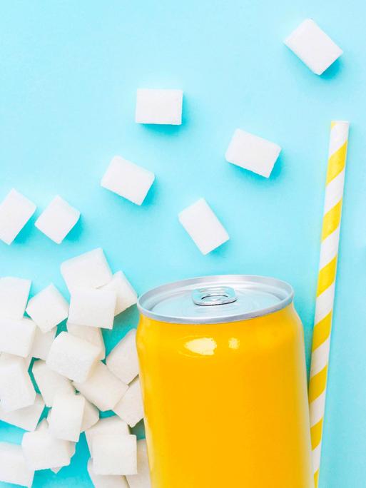 Gelbe Softdrinkdose auf hellblauen Hintergrund. Um die Dose herum liegen Würfelzucker und ein gelb-weißer Strohhalm.