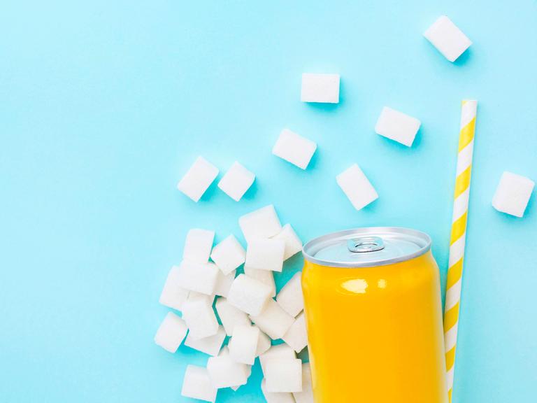 Gelbe Softdrinkdose auf hellblauen Hintergrund. Um die Dose herum liegen Würfelzucker und ein gelb-weißer Strohhalm.