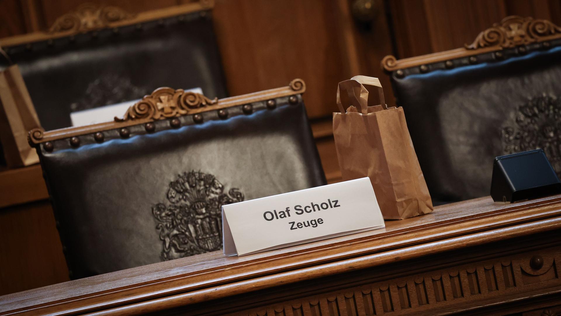 Ein Plastikschild mit dem Text "Olaf Scholz Zeuge" steht auf einem Platz auf der Senatsbank.