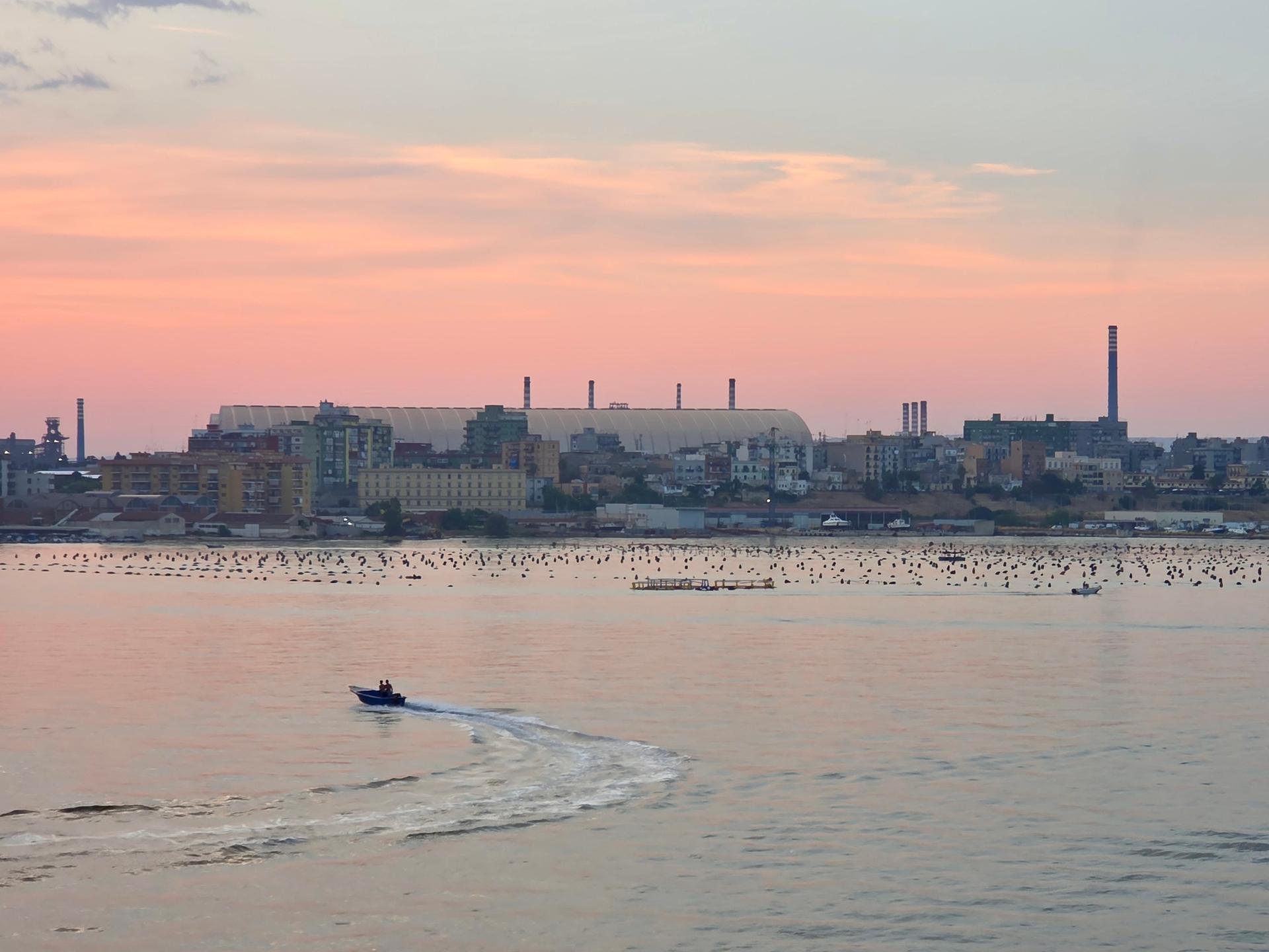 Die Kuppel des Stahlwerks in Tarent, davor Häuser und das Meer im Abendrot.