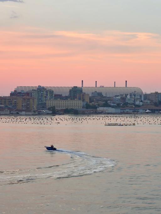 Die Kuppel des Stahlwerks in Tarent, davor Häuser und das Meer im Abendrot.