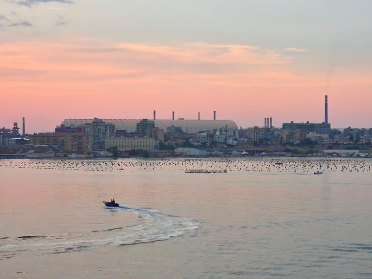 Die Kuppel des Stahlwerks in Tarent, davor Häuser und das Meer im Abendrot.