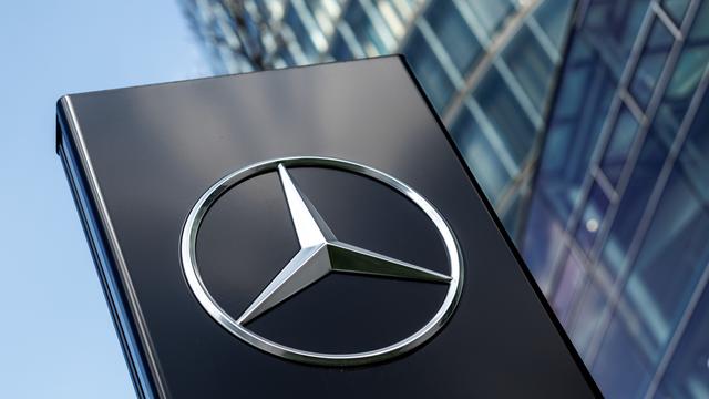 Ein silberner Mercdes-Benz-Stern prangt auf einem schwarzen Untergrund. Im Hintergrund ein Bürogebäude