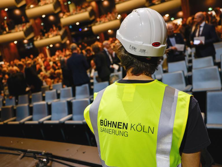 Ein als Bauarbeiter verkleideter Schauspieler sitzt auf der Bühne. Oper und Schauspiel Köln feiern den Wiedereinzug in ihre angestammten Häuser am Offenbachplatz mit einer großen, festlichen Gala. Die Sanierung der Kölner Bühnen hat 14 Jahre gedauert.