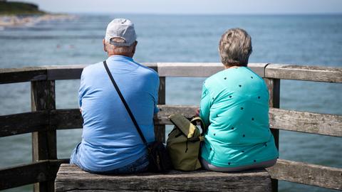 Zwei ältere Menschen blicken von der Seebrücke Graal-Müritz auf die Ostsee. Sie sind nur von hinten zu sehen. Er trägt eine Mütze, zwischen ihnen steht eine Tasche. 