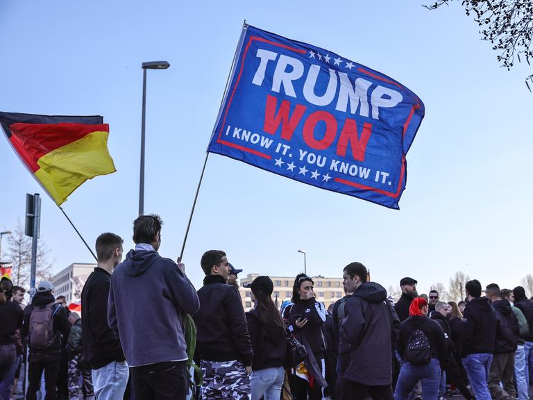 Rechtsextreme Aktivisten der AfD halten ein Transparent zur Unterstützung von US-Präsident Donald Trump hoch, bei einer Demonstration im März 2025 in Berlin, daneben schwingt eine Deutschlandfahne.