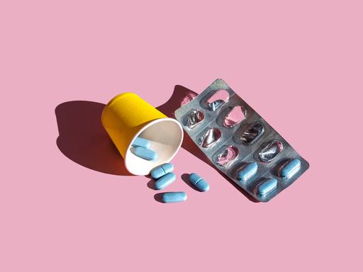 Ein Blister mit blauen Pillen liegt neben einem gelben Becher auf rosa Grund. Vor dem Becher liegen ein paar lose Pillen.