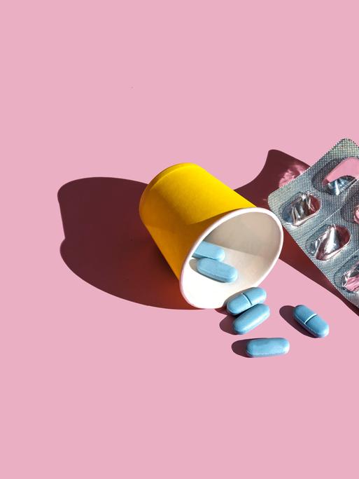 Ein Blister mit blauen Pillen liegt neben einem gelben Becher auf rosa Grund. Vor dem Becher liegen ein paar lose Pillen.
