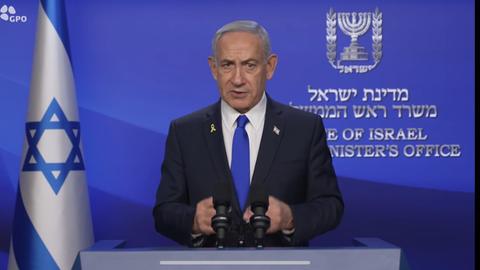 Netanjahu steht vor einer blauen Wand mit weißer hebräischer Schrift und neben einer israelischen Flagge. Er sagt etwas und gestikuliert dazu mit beiden Händen. 