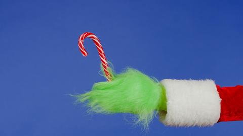 Die grüne Fellhand des Grinch in einem Weihnachtsmann-Mantel hält eine Zuckerstange.
