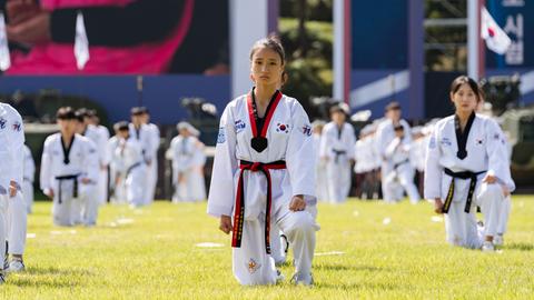 Bei einer großen Veranstaltung unter freiem Himmel sind Taekwondo-Kämpfer aufgestellt. Eine junge Frau in ihrem weißen Kampfanzug schaut in die Kamera. Bei einer großen Veranstaltung unter freiem Himmel sind Taekwondo-Kämpfer aufgestellt. Eine junge Frau in ihrem weißen Kampfanzug schaut in die Kamera.