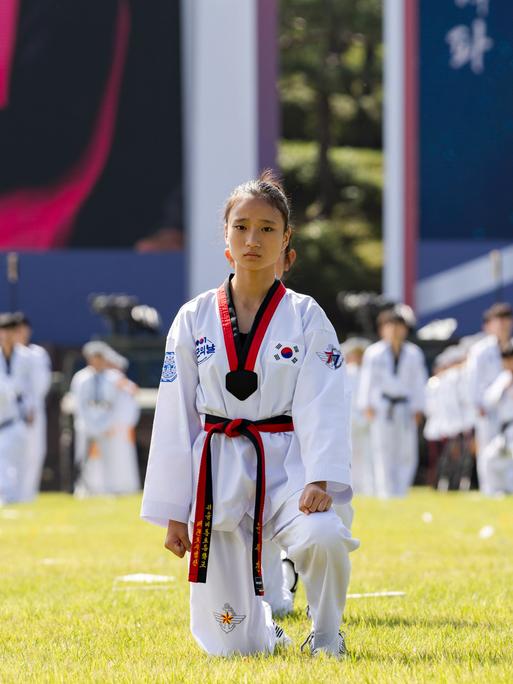 Bei einer großen Veranstaltung unter freiem Himmel sind Taekwondo-Kämpfer aufgestellt. Eine junge Frau in ihrem weißen Kampfanzug schaut in die Kamera.
