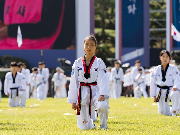 Bei einer großen Veranstaltung unter freiem Himmel sind Taekwondo-Kämpfer aufgestellt. Eine junge Frau in ihrem weißen Kampfanzug schaut in die Kamera.