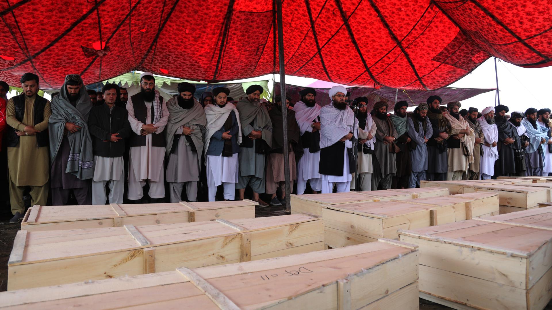 Unter einem roten Zeltdach stehen trauernde Männer an mehreren Holzsärgen.