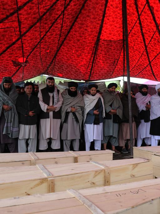 Unter einem roten Zeltdach stehen trauernde Männer an mehreren Holzsärgen.