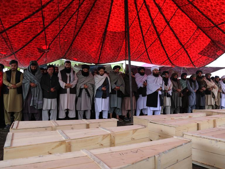 Unter einem roten Zeltdach stehen trauernde Männer an mehreren Holzsärgen.