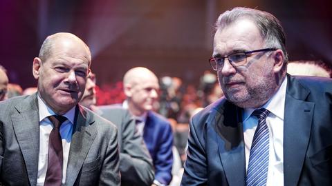 Bundeskanzler Olaf Scholz und BDI-Chef Siegfried Russwurm sitzen vor weiteren Personen in einer Veranstaltungshalle.