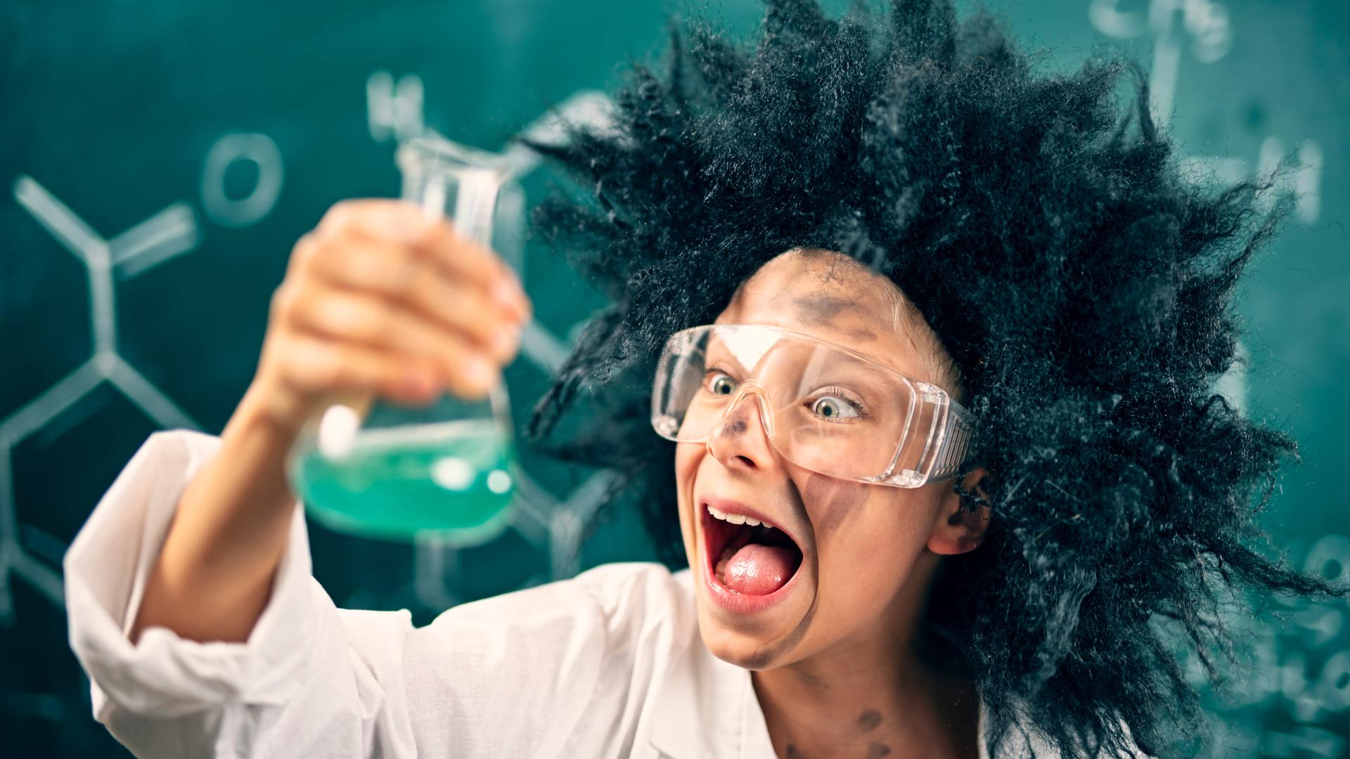 Ein als "verrückter Wissenschaftler" verkleidetes Kind mit Laborbrille steht vor einer Tafel und schaut begeistert auf ein Laborgefäß in seiner Hand, das mit einer grünen Flüssigkeit gefüllt ist. 