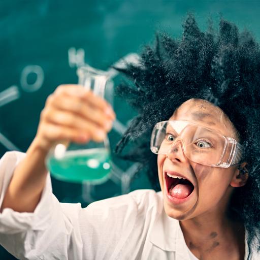 Ein als "verrückter Wissenschaftler" verkleidetes Kind mit Laborbrille steht vor einer Tafel und schaut begeistert auf ein Laborgefäß in seiner Hand, das mit einer grünen Flüssigkeit gefüllt ist. 