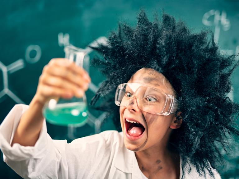 Ein als "verrückter Wissenschaftler" verkleidetes Kind mit Laborbrille steht vor einer Tafel und schaut begeistert auf ein Laborgefäß in seiner Hand, das mit einer grünen Flüssigkeit gefüllt ist. 