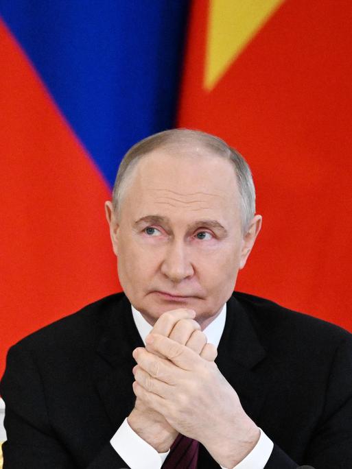Russlands Präsident Wladimir Putin sitzt vor roten Fahnen und reibt sich die Hände.