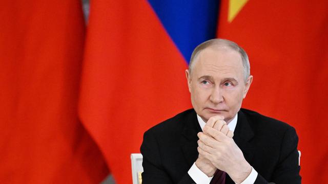 Russlands Präsident Wladimir Putin sitzt vor roten Fahnen und reibt sich die Hände.