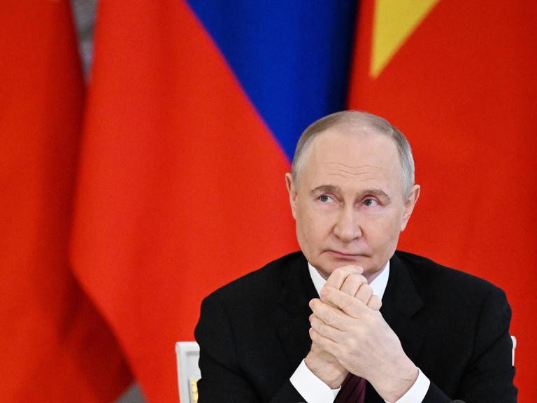 Russlands Präsident Wladimir Putin sitzt vor roten Fahnen und reibt sich die Hände.