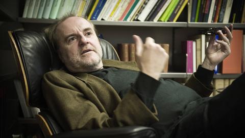 Peter Schneider sitzt in seiner Praxis bequem in einem Lehnsessel, hinter sich ein gut gefülltes Bücherregal.