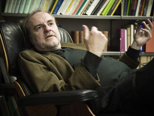 Peter Schneider sitzt in seiner Praxis bequem in einem Lehnsessel, hinter sich ein gut gefülltes Bücherregal.