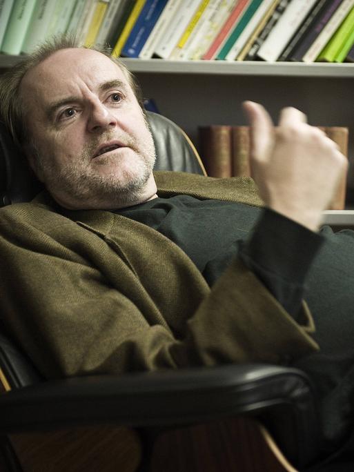 Peter Schneider sitzt in seiner Praxis bequem in einem Lehnsessel, hinter sich ein gut gefülltes Bücherregal.