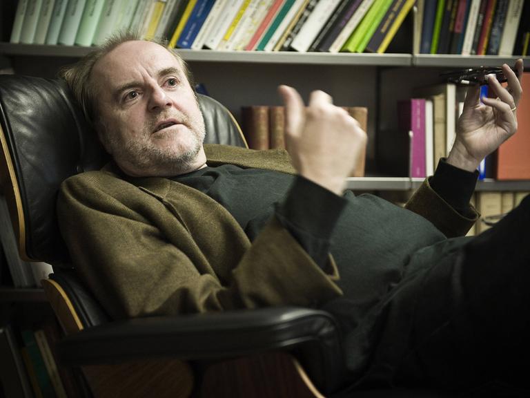 Peter Schneider sitzt in seiner Praxis bequem in einem Lehnsessel, hinter sich ein gut gefülltes Bücherregal.