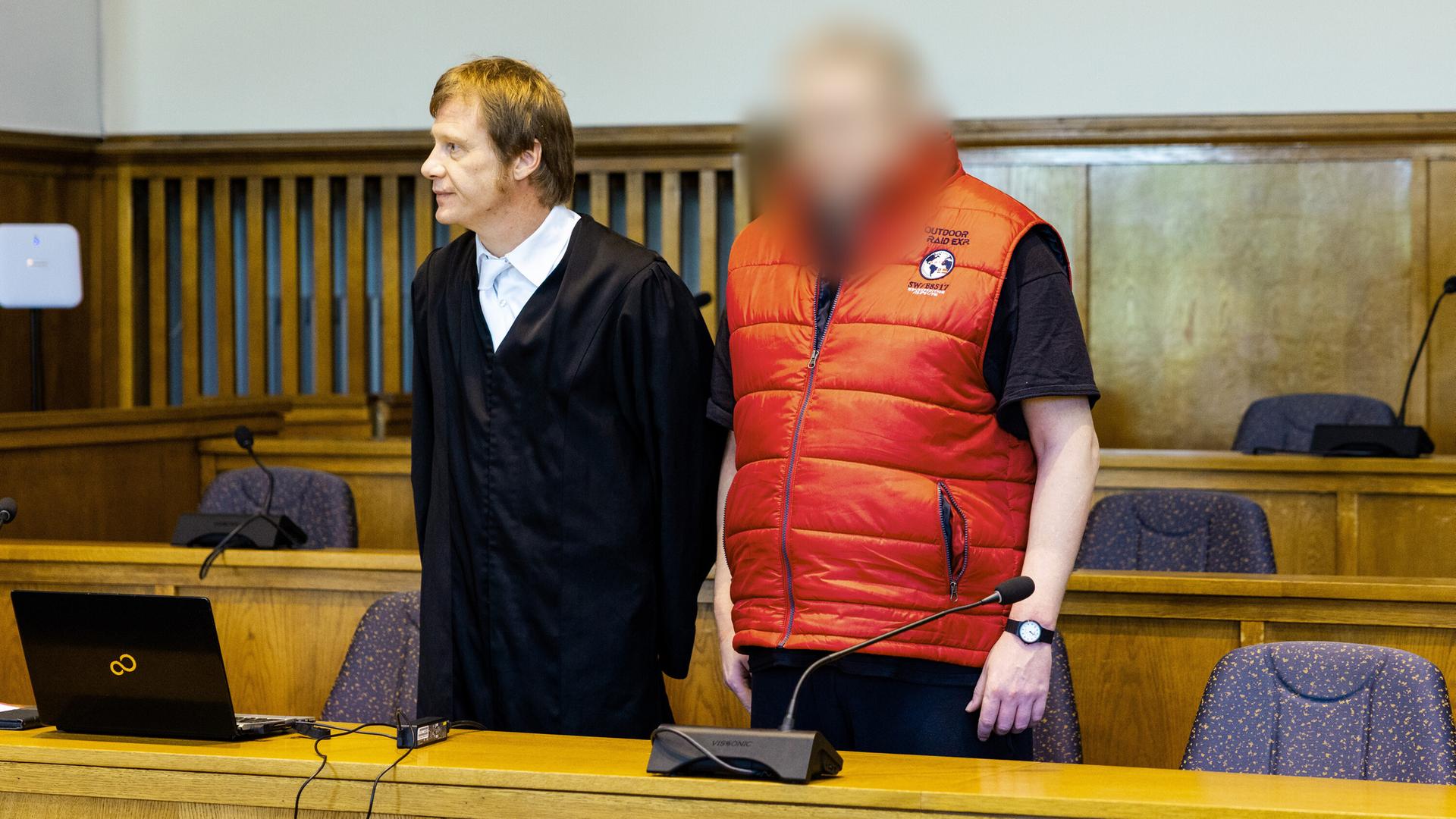 Die beiden stehen nebeneinander hinter einem Tisch. Der Anwalt trägt schwarz, der Angeklagte eine ärmellose rote Jacke. Sein Gesicht ist verpixelt. Die beiden stehen nebeneinander hinter einem Tisch. Der Anwalt trägt schwarz, der Angeklagte eine ärmellose rote Jacke. Sein Gesicht ist verpixelt.