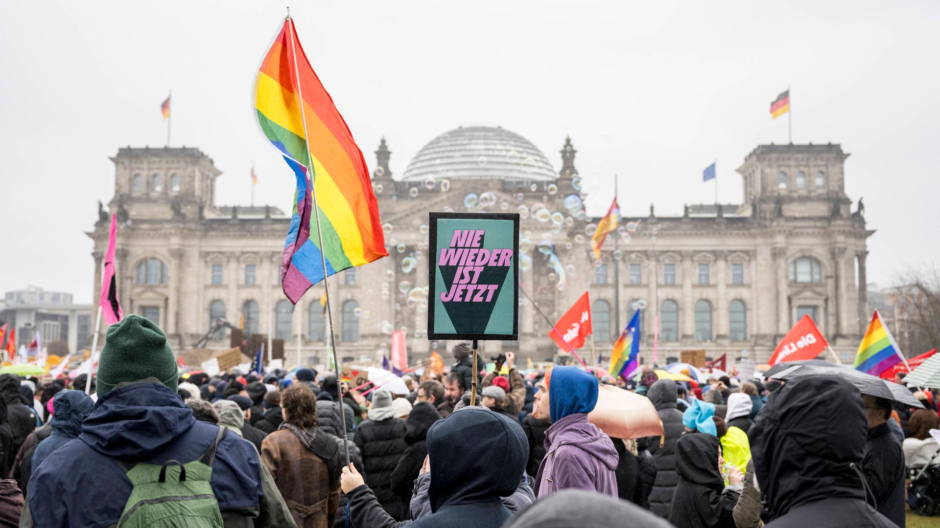 Demonstrierende schwenken Regenbogenflaggen und andere Fahnen vor dem Reichstagsgebäude in Berlin. Auf einem Schild steht er Slogan "Nie wieder ist jetzt".